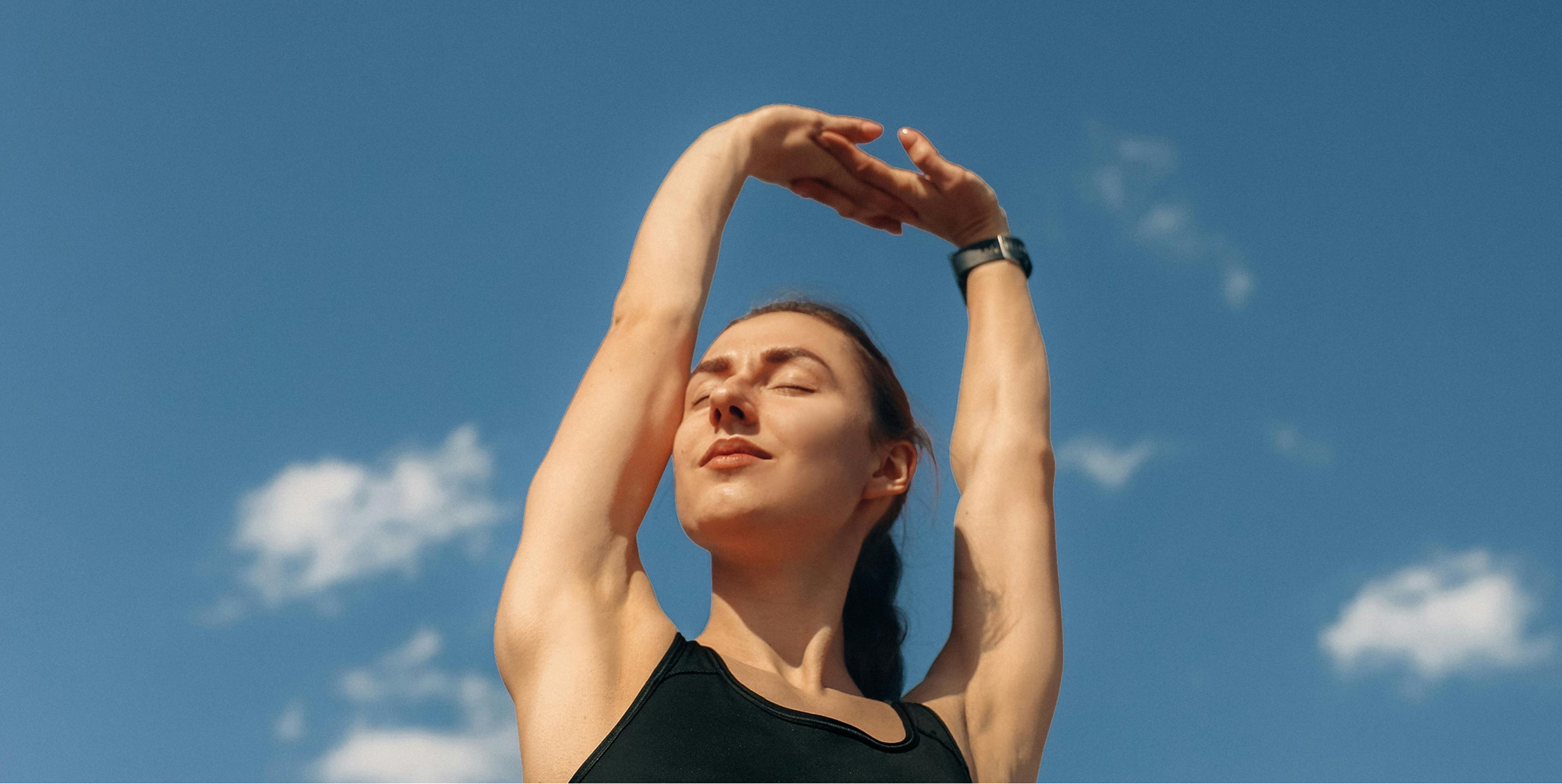 Woman stretching outdoors under a clear blue sky, wearing a black athletic top and fitness tracker